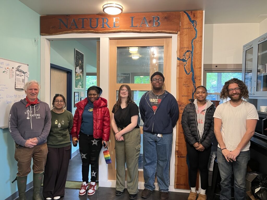 6 people (high school students and mentors of a range of ages, ethnicities, genders) stand smiling in front of door that has a wide wood frame that reads "NATURE Lab" in decorative text.